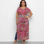 A model wearing a plus-size short-sleeve top in a vibrant fuchsia and multicolor abstract print. The sheer power mesh blouse features a round neck and side slits, and is styled with a statement silver belt at the waist.
