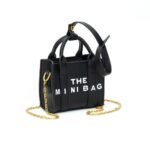 A compact, structured black faux leather mini bag featuring a prominent white text message design. The bag includes a small top handle, top zipper closure, a short chain strap for bag-charm attachment, and a long detachable shoulder strap.