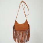 A crescent-shaped suede crossbody bag in a soft neutral tone, featuring long bohemian fringe trim, an adjustable shoulder strap, and mixed metal hardware.